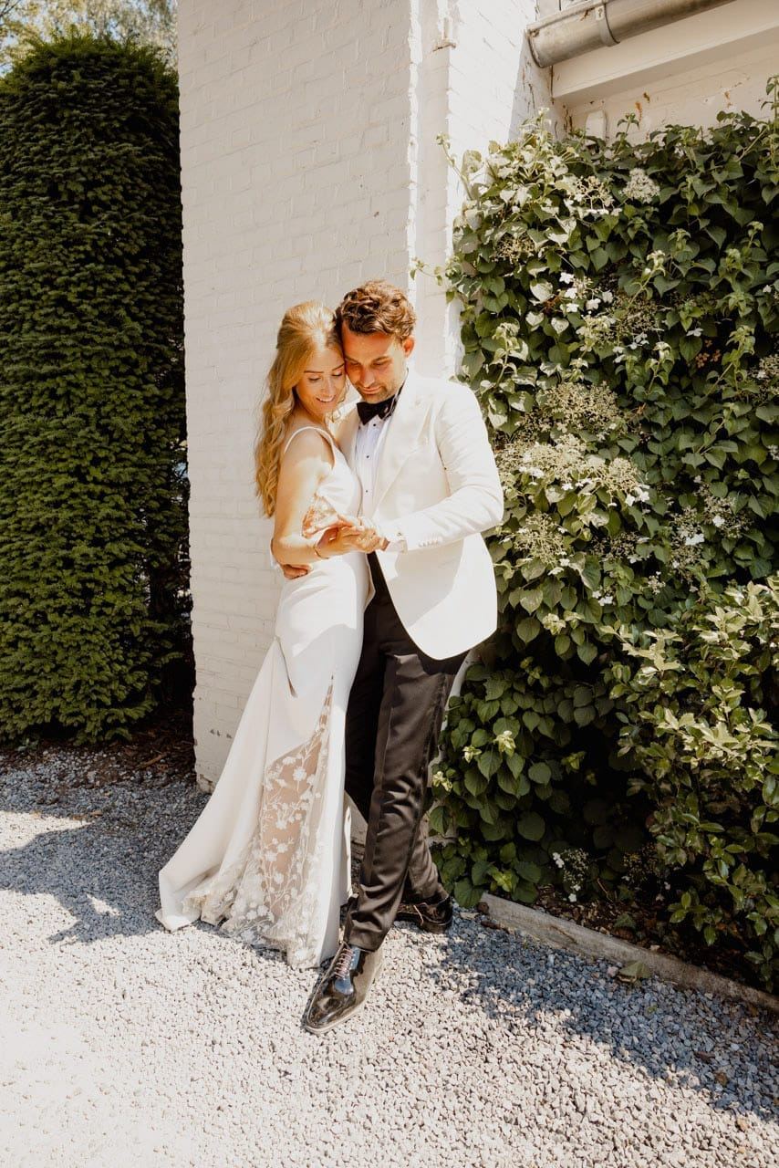 Couple in a white tuxedo and a wedding dress, close together against a white wall and climbing plants